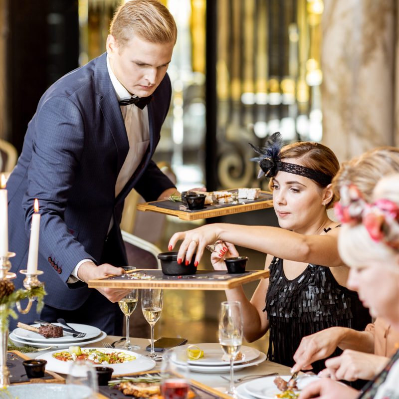 Elegantly dressed group of people having a festive dinner during New Year Eve at the luxury restaurant, waiter serving dishes